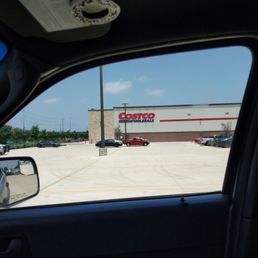 The image captures a view from inside a vehicle looking out through the passenger side window at a Costco Wholesale store. The parking lot is mostly empty with a few parked cars, and the store's sign is visible on the building. The sky is clear with some clouds, and there is a pole in the foreground. The vehicle's side mirror and part of the interior are also visible.