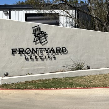 Frontyard Brewing - Front entrance - The image shows the front exterior of Frontyard Brewing, with a white textured wall bearing the business logo, which includes a black chair graphic and the name 'FRONTYARD BREWING' in bold black letters. The wall is set against a background of trees and a building with a black vent or window. The ground in front of the wall is a paved sidewalk with some grass and small plants nearby. - Breweries Near Me - Spicewood, Texas
