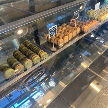 Black Lion Cafe - Dessert - The image shows a glass display case filled with an assortment of macarons arranged in rows. The macarons are in different colors, including green, brown, and orange, indicating various flavors. Small signs with labels are placed among the macarons, providing flavor descriptions. The case is part of a café or bakery setting, with reflections of the interior visible on the glass surface. - Cafes Near Me - Silver Spring, Maryland