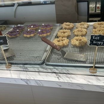 Mochi Ring - The image shows two metal trays on a marble countertop, each containing different types of snacks. The left tray has purple ube-flavored mochi donuts decorated with small white toppings, while the right tray holds rice krispy-style Korean corn dogs with a crunchy coating. Small chalkboard signs label the items as 'Ube' and 'Rice Krispy.' The background includes part of the store interior, with a person partially visible on the right. - Donuts Near Me - Wauwatosa, Wisconsin