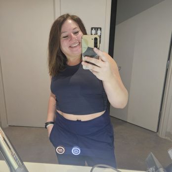 Sweathouz - Great "getting ready" stations! - A woman with shoulder-length brown hair is taking a mirror selfie in a restroom, smiling, wearing a black sleeveless top and dark pants, with her left hand in her pocket. - Saunas Near Me - Chicago, Illinois
