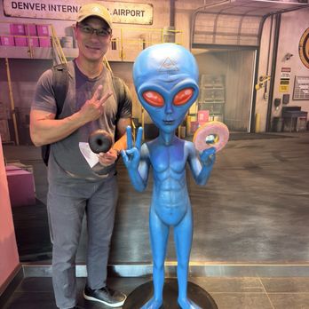 Voodoo Doughnut - Voodoo donuts - A man stands beside a blue alien statue holding a donut and a black ball, making a peace sign at Denver International Airport. - Donuts Near Me - Denver, Colorado