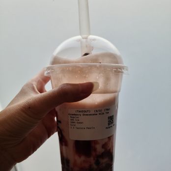 Palgong Tea - The drink I ordered - A hand is holding a large clear plastic cup filled with strawberry cheesecake milk tea containing tapioca pearls at the bottom. The cup has a domed lid with a straw inserted through it. A label on the cup indicates the order details, including the drink type, size, and ingredients. The background is plain and neutral, emphasizing the beverage. - Bubble Tea Near Me - Thornhill, Ontario