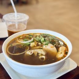 Bun Bo Hue - A bowl of bun bo hue, a traditional Vietnamese spicy beef noodle soup, contains broth, herbs, sliced onions, eggs, and meat, served on a white tray. A plastic cup with iced coffee and a straw is placed behind the bowl. The setting appears to be inside a restaurant with a blurred background.
