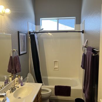 Mk Repairs - Guest Bath before picture. - A small bathroom features a bathtub with a black shower curtain, a toilet, and a double-sink vanity with purple towels hanging on the wall. There is a window above the shower area allowing natural light, and the walls are painted gray. The floor has wood-like flooring, and a black trash bin is placed near the toilet. The bathroom includes a mirror, framed artwork, and various toiletries on the sink counter. - General Contractors Near Me