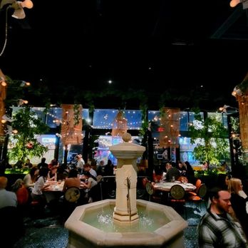ilili DC - The image shows a restaurant interior with a central stone fountain, surrounded by tables filled with diners. The space is decorated with hanging light fixtures resembling birdcages, greenery, and large windows in the background. The setting appears lively and well-lit, with a mix of natural and artificial lighting, and a dark ceiling contrasting with the bright decor. - Lebanese Near Me - Washington, DC, District of Columbia