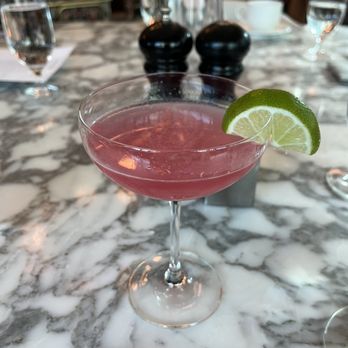 fl.2 - Cosmopolitan - A pink cocktail served in a coupe glass with a lime wedge garnish is positioned on a marble table. The background includes water glasses, a white cup, and dark condiment shakers, indicating a restaurant environment with a modern and elegant interior. - New American Near Me - Pittsburgh, Pennsylvania