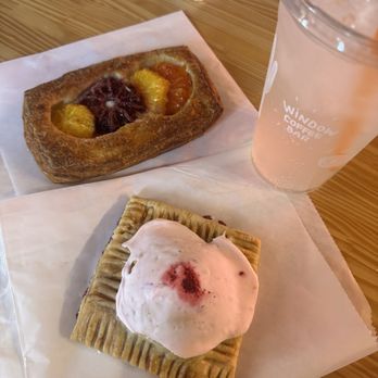 Window Coffee Bar - Raspberry prickly pear fruit tart Fruit danish Mango coconut lotus energy - The image shows a wooden table with two pastries and a beverage at Window Coffee Bar in Phoenix, AZ. The first pastry is a fruit Danish with a rectangular shape, topped with colorful fruit pieces including pineapple, orange, and a dark berry. The second pastry is a square with a pink frosting or glaze and a small red topping. A clear plastic cup with a pink lemonade or similar drink, labeled 'Window Coffee Bar,' is positioned to the right of the pastries. The table surface is light-colored wood, and a white napkin is partially visible in the background. - Coffee & Tea Near Me - Phoenix, Arizona