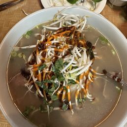 A large bowl of Vietnamese pho is filled with clear broth, bean sprouts, and herbs, topped with a drizzle of dark sauce. Surrounding the bowl are side plates containing lime wedges, basil leaves, and additional bean sprouts, along with a small bowl of noodles. The setting is on a wooden table with utensils and a glass of water nearby, indicating a casual dining environment.