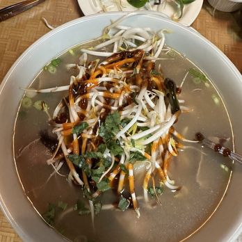 Pho King Orem - A large bowl of Vietnamese pho is filled with clear broth, bean sprouts, and herbs, topped with a drizzle of dark sauce. Surrounding the bowl are side plates containing lime wedges, basil leaves, and additional bean sprouts, along with a small bowl of noodles. The setting is on a wooden table with utensils and a glass of water nearby, indicating a casual dining environment. - Vietnamese Near Me - Orem, Utah
