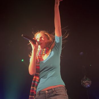The Sinclair - A woman with red hair is performing on stage, holding a microphone close to her mouth. She is wearing a light blue shirt and a colorful striped scarf. Her right arm is raised high above her head, and stage lights illuminate her from below, creating a dramatic effect. The background is dark with some stage equipment and a disco ball visible. - Music Venues Near Me - Cambridge, Massachusetts