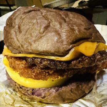 Cebu Chicago - Ube Chorizo Sandwich - A burger with a purple sweet potato bun, fried chicken patty, melted cheese, and other ingredients, placed on crinkled paper. The bun appears soft and vibrant, with the fried chicken and cheese visible between the buns. The background shows a casual setting. - Filipino Near Me - Chicago, Illinois