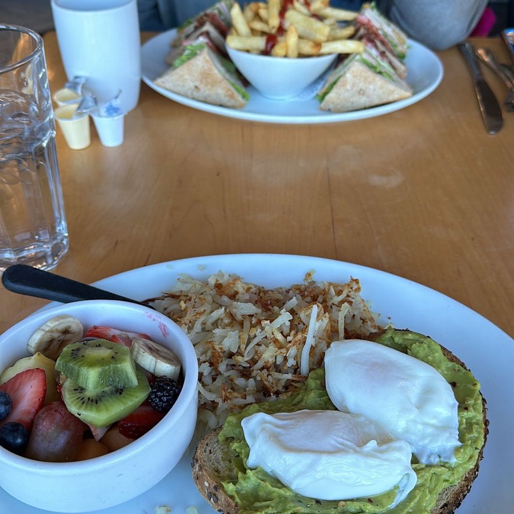 Poached eggs avocado toast with fruit in foreground. Triple decker club sandwich with fries in background. Poached eggs avocado toast with fruit in foreground. Triple decker club sandwich with fries in background.