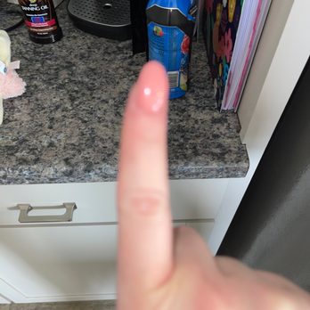 JL Nails - A finger is extended towards a kitchen countertop that has a bottle of tanning oil, a coffee machine, and a colorful notebook or folder. - Nail Salons Near Me - Howell, Michigan