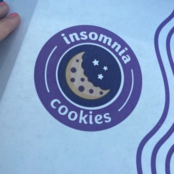 Insomnia Cookies - Round two! - The image shows a paper bag with the Insomnia Cookies logo printed on it. The logo features a stylized crescent moon with a face, surrounded by three stars, all within a purple circular background. The words "insomnia" and "cookies" are written in white, curved around the circle. The bag appears to be held by a person with a hand visible on the left side of the image. The background is dark, and part of the bag's design includes purple wavy lines on the right side. - Desserts Near Me - Albuquerque, New Mexico