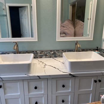 ASR Construction - Clean, coastal vibe in our updated space - The image shows a bathroom vanity with a white marble countertop that has dark veining. Two rectangular vessel sinks are installed on the countertop, each with a metallic faucet. Above the sinks are two mirrors framed in white, reflecting parts of the room. The backsplash consists of small, rectangular, multicolored tiles. The cabinetry below is white with black knobs, and the wall behind the mirrors is painted in a light, muted color. - General Contractors Near Me