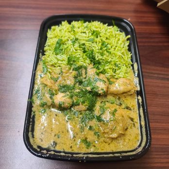 Xpression - Cream chicken - A black plastic container on a wooden table contains a serving of bright green rice and pieces of chicken in a creamy sauce with herbs. The rice appears fluffy and is separated from the chicken and sauce, which is garnished with chopped green herbs. The setting suggests a takeout meal, possibly from an Indian fusion restaurant. - Indian Near Me - Berkeley, California