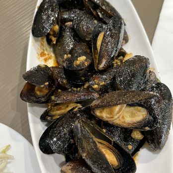 Pho Saigon 75 - A white oval plate contains cooked mussels with dark shells, some open revealing the meat inside. The mussels are seasoned with garlic, spices, and possibly chili flakes, with visible bits of garlic and seasoning on and around them. The dish appears to be a seafood appetizer or main course, presented on a neutral-colored surface. - Vietnamese Near Me - Fairfax, Virginia