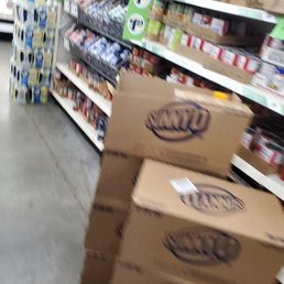 A grocery store aisle features shelves filled with packaged food items, including bread and snacks. Several large cardboard boxes are stacked in the foreground, partially obstructing the view of the shelves. The store has a concrete floor, and the image appears slightly blurry.