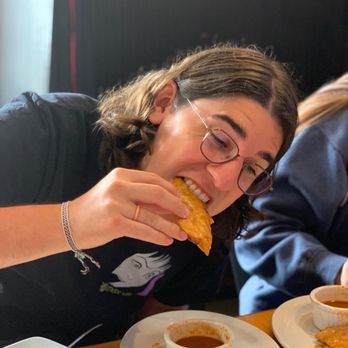 D’ Lua Pancake & Burrito House - Quesabirrias with their delicious consome soup - A woman with glasses and shoulder-length hair is holding a taco and smiling at a restaurant table with various dishes and sauces. - Mexican Near Me - Moline, Illinois