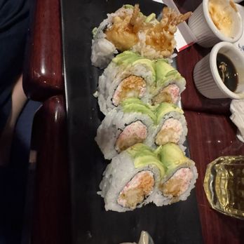 Shibui - A black rectangular serving platter holds multiple sushi rolls, including avocado-topped rolls and shrimp tempura rolls. Accompanying the sushi are small cups of soy sauce, wasabi, and pickled ginger. The platter is placed on a dark wooden table, with additional cups and a glass of beverage visible in the background. - Japanese Near Me - Sunset, Florida