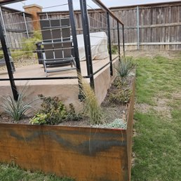 A backyard patio features a black metal railing, outdoor furniture including a chair and a covered grill, with a garden bed containing various plants.