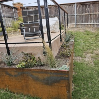 Austin Creative Landscaping - A backyard patio features a black metal railing, outdoor furniture including a chair and a covered grill, with a garden bed containing various plants. - Landscaping Near Me - Austin, Texas