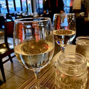 Flavio Restaurant DC - Two glasses of white wine are placed on a restaurant table, with a blurred background showing other diners and a person standing. - Italian Near Me - Washington, DC, District of Columbia