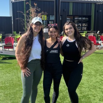 The Foundry - Three women pose together outdoors on a grassy area with a building and string lights in the background, smiling at the camera. - Yoga Near Me - Avondale, Arizona
