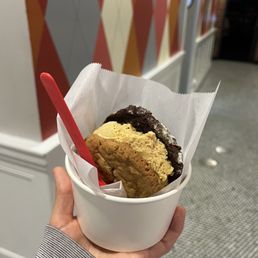 Chocolate Chip Ice Cream Sandwich - A person is holding a white paper-lined cup with two ice cream sandwiches, one with a chocolate cookie and the other with a vanilla cookie, in an indoor setting. The background features a colorful, geometric-patterned wall and part of a brick wall with a fireplace. The person's hand is visible, wearing a gray and white striped sleeve.