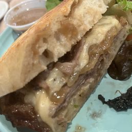 Short Rib " Panini - A sandwich featuring melted cheese, sliced meat, and bread is presented on a light blue plate. The sandwich is partially cut, revealing the ingredients inside. A side salad with mixed greens and a small container of dipping sauce are also visible in the background. The image captures the details of the sandwich's texture and ingredients, emphasizing its hearty appearance.