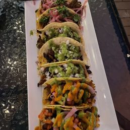 Carne Asada - Six tacos with different toppings, including guacamole, pickled onions, and shredded vegetables, served on a white rectangular plate at a restaurant.