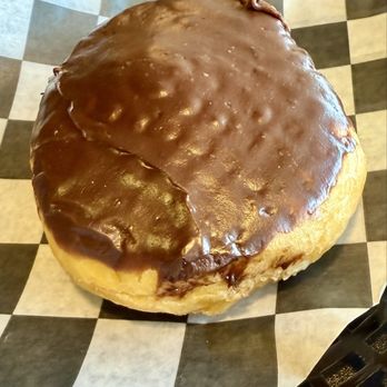 Jack's Donuts - Boston Cream - yum - A sizable donut with a glossy chocolate glaze covers the top, resting on black and white checkered paper, likely from a donut shop. - Donuts Near Me - Greenfield, Indiana