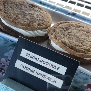 Sweetspot - Jumbo Snickerdoodle Cookie Sandwich. - Two snickerdoodle cookies with a generous layer of white cream filling are placed on a tray. The cookies are large, round, and have a textured surface typical of snickerdoodles. A small sign in front of the tray labels them as 'Snickerdoodle Cookie Sandwich.' The background includes a reflective surface and part of a decorative tablecloth, indicating an indoor bakery or cafe environment. - Cupcakes Near Me - Redding, California