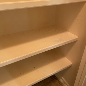 Texas Cleaning Services - Pantry shelves were not cleaned - A set of four beige pantry shelves is depicted, with visible dust, dirt, and signs of neglect. The shelves are mounted on a wall, with the bottom shelf showing more dirt and some discoloration. The surrounding area includes a section of a wall and a tiled floor, suggesting an indoor storage space that has not been cleaned or maintained recently. - Home Cleaning Near Me - Austin, Texas