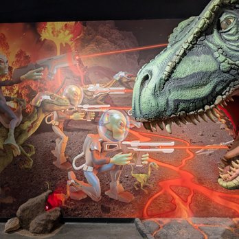 Shell - An animated scene shows aliens riding a dinosaur and shooting laser guns at a large dinosaur with an open mouth, set against a volcanic background. - Gas Stations Near Me - Dragoon, Arizona