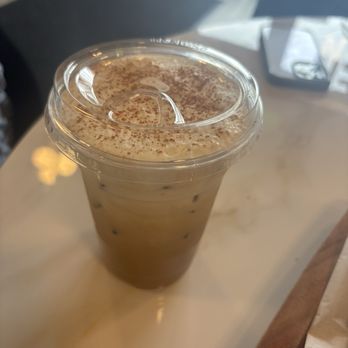 Lore’s Cafe - A clear plastic cup filled with iced coffee topped with whipped cream and cocoa powder, placed on a light-colored table. - Cafes Near Me - Beacon, New York