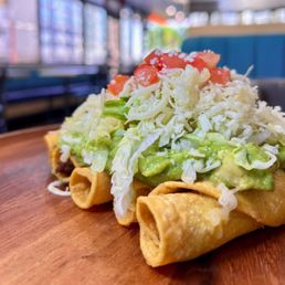 Rolled Tacos - A rolled taco with shredded cheese, lettuce, guacamole, and diced tomatoes on top, served on a wooden plate in a restaurant setting.