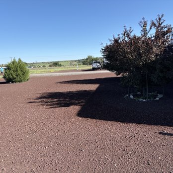 Springerville RV Park - Typical RV space - A gravel parking lot features bushes and a large vehicle parked on the right side. The area is open with a clear blue sky overhead and distant landscape visible in the background. Shadows from the bushes are cast on the ground, and there are a few cars and structures in the distance. - RV Parks Near Me - Springerville, Arizona