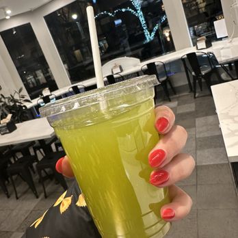 Taboonette - a woman holding a glass of green juice - A person with red-painted nails holds a large clear plastic cup filled with a green juice and a white straw. The background shows a cafe interior with white tables, black chairs, and large windows reflecting a tree with blue string lights outside. The cafe has a modern decor with bright lighting and a clean, minimalistic design. - Mediterranean Near Me - Brooklyn, New York