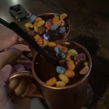 The Copper Cup - Trix Cereal - Two copper mugs contain a dark drink topped with colorful cereal pieces, held by a person in a dimly lit setting. - Cocktail Bars Near Me - Des Moines, Iowa