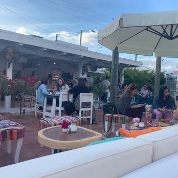 Tayrona - The image shows an outdoor dining area at a Colombian-themed restaurant or bar, with several groups of people seated at tables and on cushioned benches. The space features colorful, woven stools, potted plants, and large umbrellas providing shade. In the background, a bar area is visible with staff serving customers. The setting appears lively and social, with a clear sky overhead and a relaxed atmosphere. - Bars Near Me - Miami, Florida