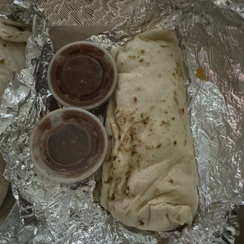 Perico's Albuquerque - smaller than my hand Chicharrones burrito large with extra meat - A burrito wrapped in foil is shown, resting on crinkled aluminum foil. Two small plastic containers filled with dark sauce are placed on top of the burrito. The background includes a textured surface and part of a paper towel roll. The image suggests a takeout or casual dining setting, with the focus on the food items. - Mexican Near Me - Albuquerque, New Mexico