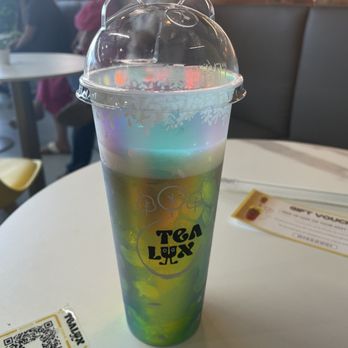 Tealux Cafe - Jasmine tea with grass jelly and no ice - A tall, rainbow-colored bubble tea with a bear-shaped plastic lid is placed on a white table inside a cafe, with customers in the background. - Bubble Tea Near Me - Ballwin, Missouri