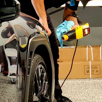 AbelTints - A man wearing a black cap and black clothing is cleaning a black vehicle with a spray bottle and cloth in an indoor setting. - Car Window Tinting Near Me - Snellville, Georgia