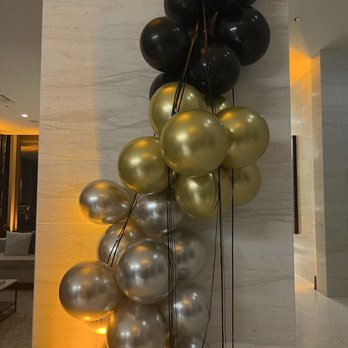 M Events - A vertical arrangement of balloons in black, gold, and silver colors is positioned against a marble wall in an indoor space. The balloons are tied with black strings, and the lighting creates reflections on their surfaces. The background includes a marble wall and part of a modern interior with warm lighting and seating areas. - Caterers Near Me