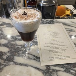 A layered coffee drink features a dark coffee base topped with a generous layer of whipped cream, dusted with cocoa powder and garnished with a small chocolate piece. The drink is served in a clear glass on a marble bar counter, with bar tools and ingredients visible in the background, indicating a bar or cafe setting.