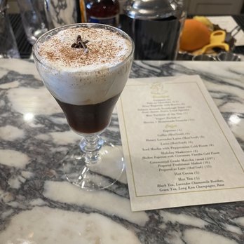 Odelia - A layered coffee drink features a dark coffee base topped with a generous layer of whipped cream, dusted with cocoa powder and garnished with a small chocolate piece. The drink is served in a clear glass on a marble bar counter, with bar tools and ingredients visible in the background, indicating a bar or cafe setting. - Coffee & Tea Near Me - Mandeville, Louisiana