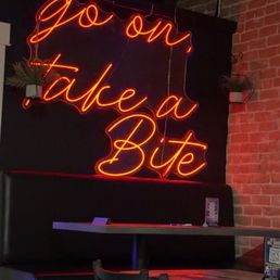 Inside - A neon sign displaying the phrase "go on take a Bite" in cursive script is mounted on a dark wall with exposed brick in an indoor dining space. The sign is illuminated in orange and red colors, and there are small potted plants on either side of the sign. The foreground shows tables and chairs arranged for seating, with some items like menus or table cards visible on the tables.