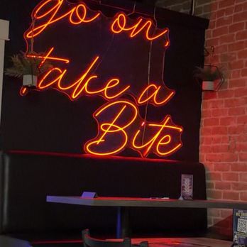 Mic Drop Comedy - Inside - A neon sign displaying the phrase "go on take a Bite" in cursive script is mounted on a dark wall with exposed brick in an indoor dining space. The sign is illuminated in orange and red colors, and there are small potted plants on either side of the sign. The foreground shows tables and chairs arranged for seating, with some items like menus or table cards visible on the tables. - Comedy Clubs Near Me - Chandler, Arizona